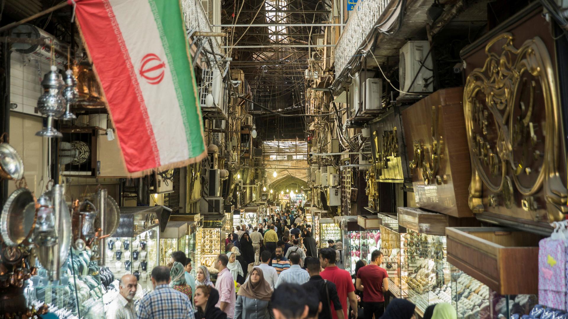 Crowds fill the bottom of the frame. On both sides are shops and the on the right side of the image is an Iranian flag