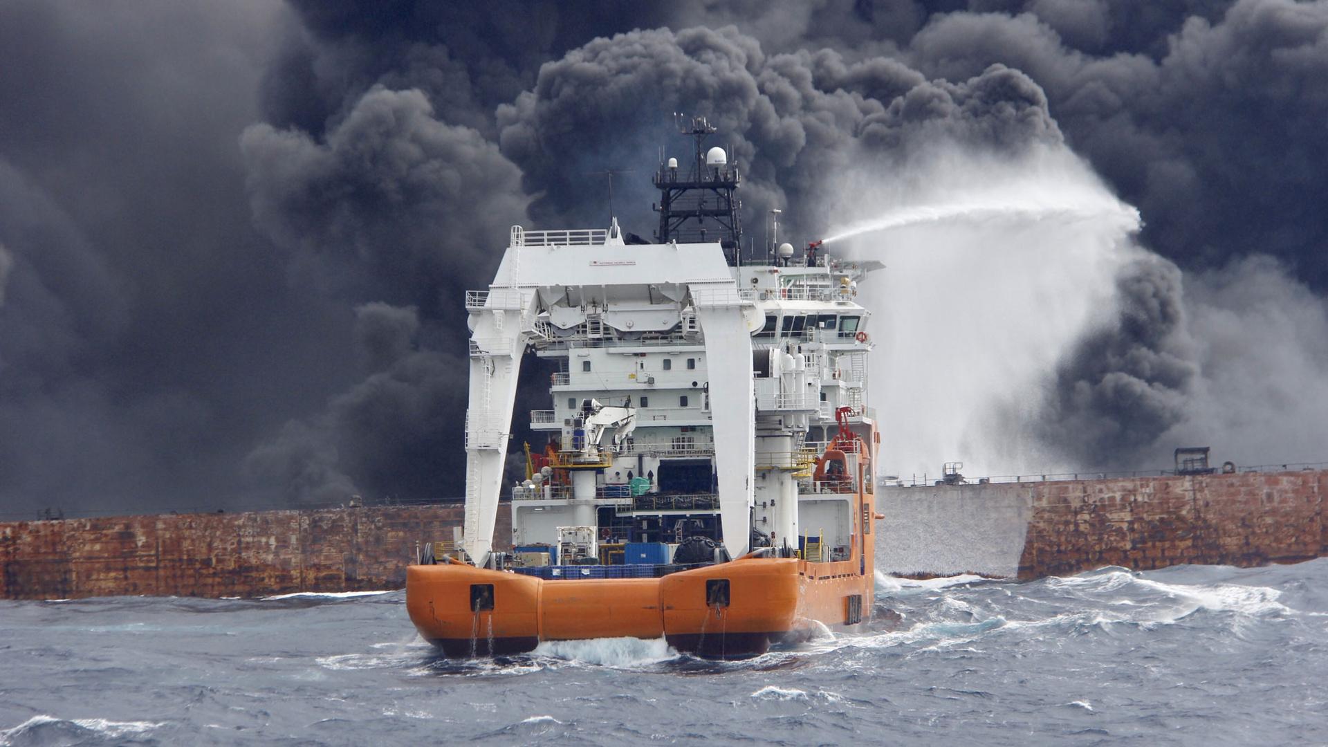 A smaller boat shoots a jet of water out a dark, billowing clouds of smoke coming from a brown metal tanker ship