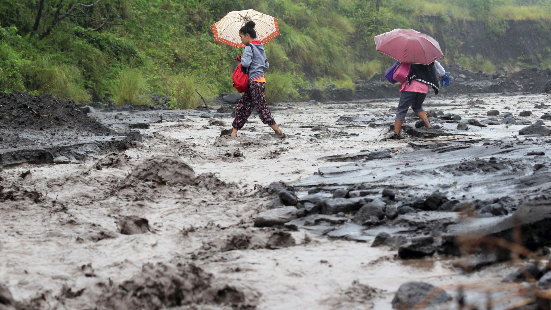 Two women hold pink and orange umbrellas as they carefully walk through a flooded area of dark earth, which is dark with volcanic ash.