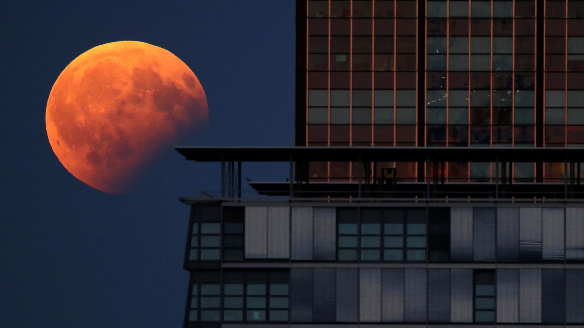 The moon is red-orange as it hangs in the sky next to the left of a high-rise building. 