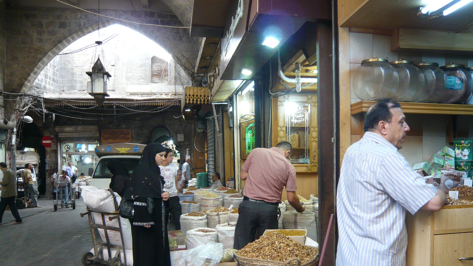 Several people look at bags of dried ingredients laid outside a shop. In the foreground, a man leans against a stall.