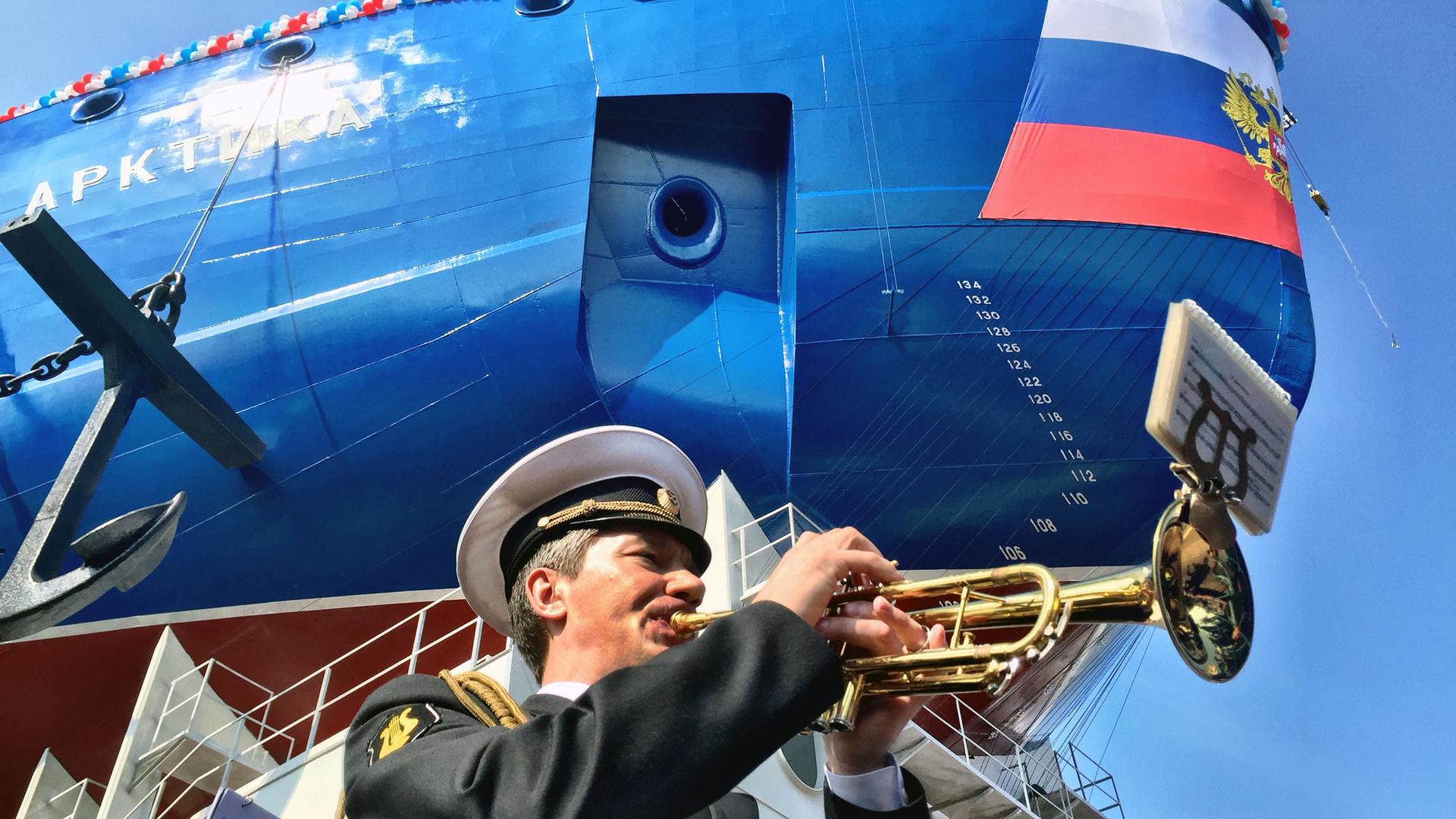 A man in a military uniform plays the trumpet. Behind him is the blue hull of the icebreaker ship Arktika.