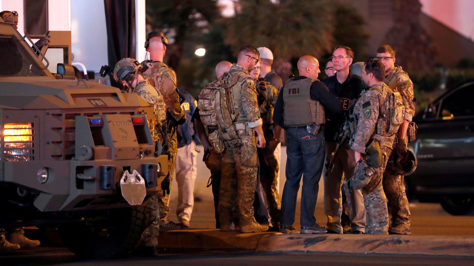 A group of men wearing camouflage clothing and FBI vests stand next to an armored car outside the Mandalay Bay Hotel in the pre-dawn light.
