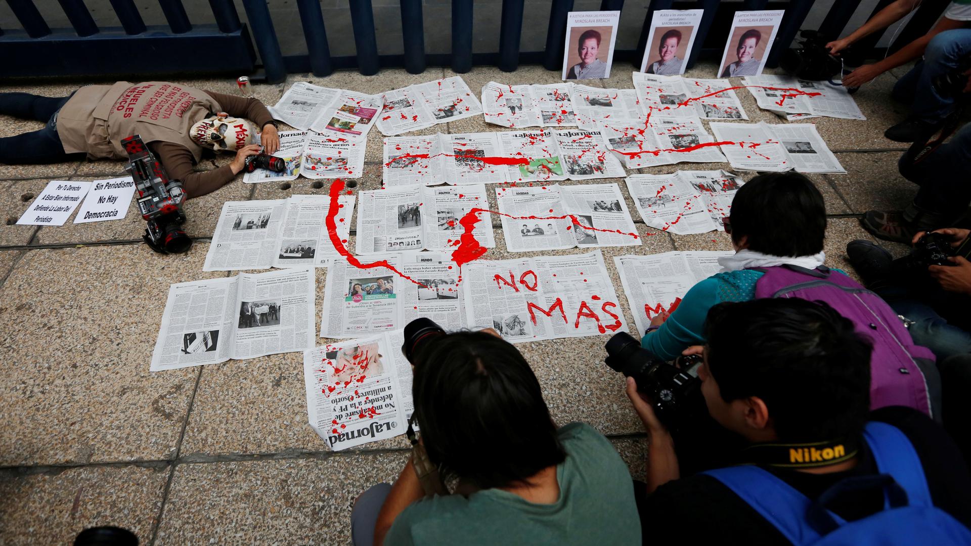 Photographers are huddled around a protest on the sidewalk. There are newspapers laid out on the sidewalk, drizzled in red paint to look like blood, while a person wearing a skull mask pretends to lay dead next to them. The words "No mas" are written.