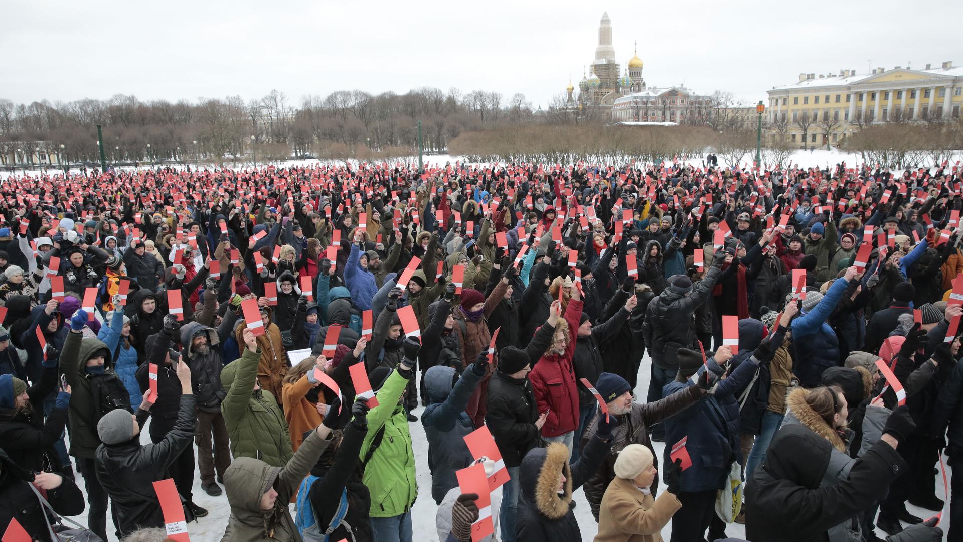 A crowd of people hold up red cards showing their support for Alexia Navalny. Behind them are some of St. Petersburg's famous landmarks. 