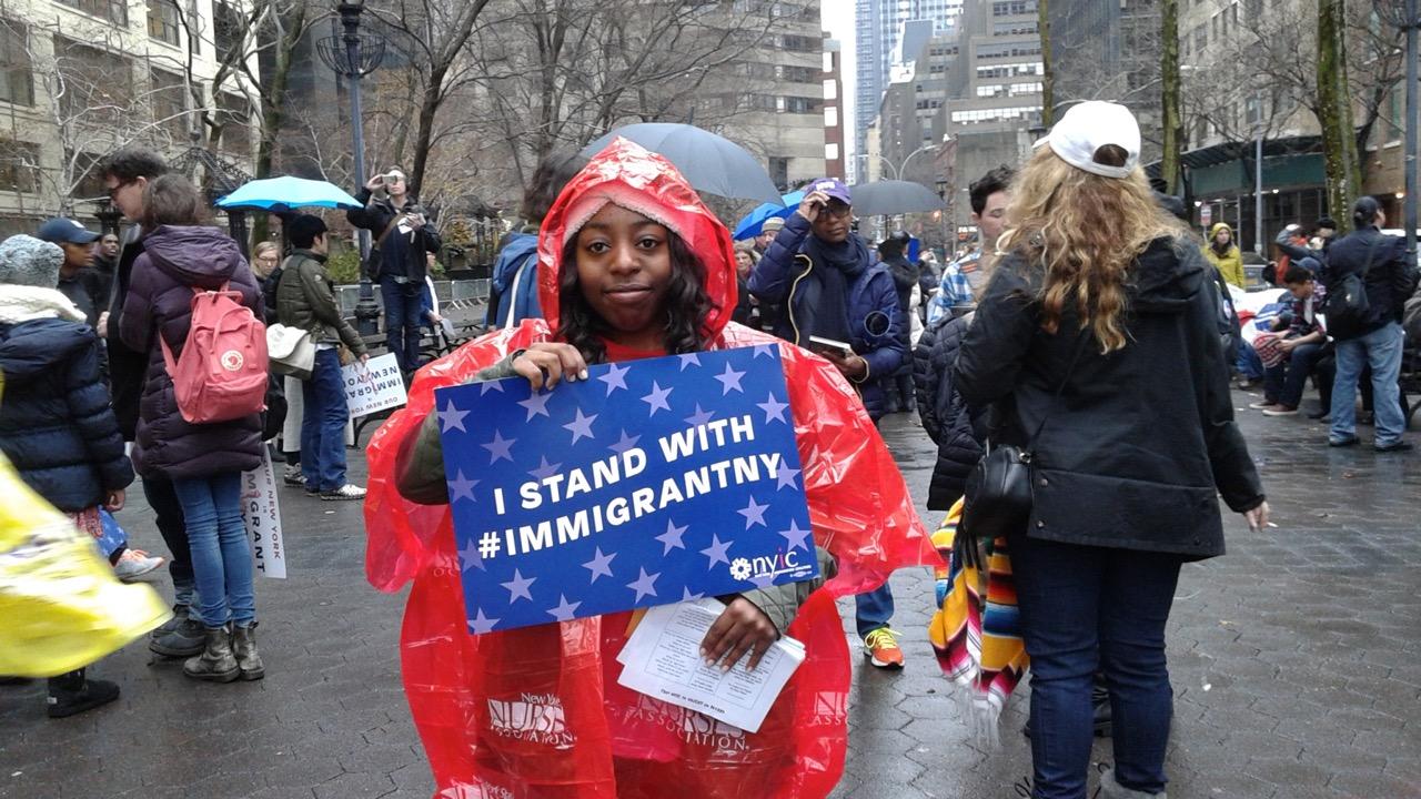 Woman in rain parka holding sign that reads "I STAND WITH #IMMIGRANTNYC"