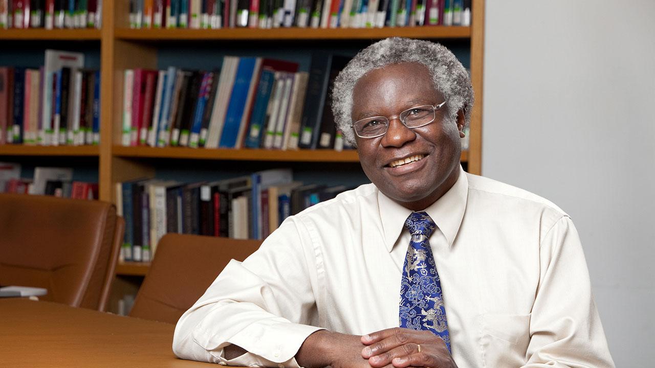 Calestous Juma poses at a table in a portrait photo. Behind him, shelves are lined with books. He has a blue tie and glasses.