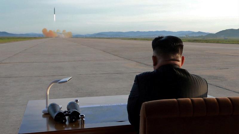 North Korean leader Kim Jong-un sits at a desk in the nearground and watches a missile launch in the distance.