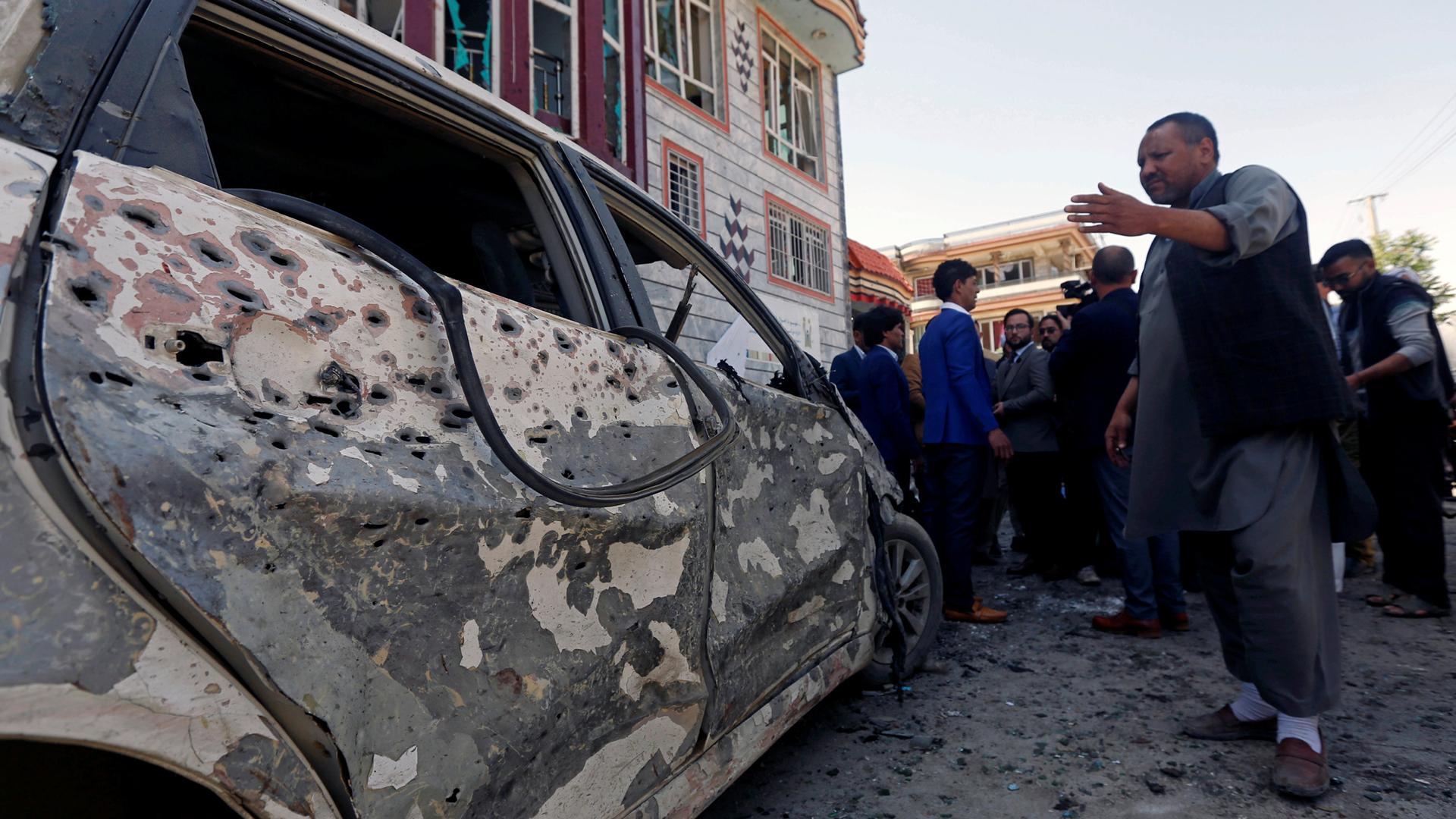 A man points left to a car that shows the impact of a bomb blast in Kabul.