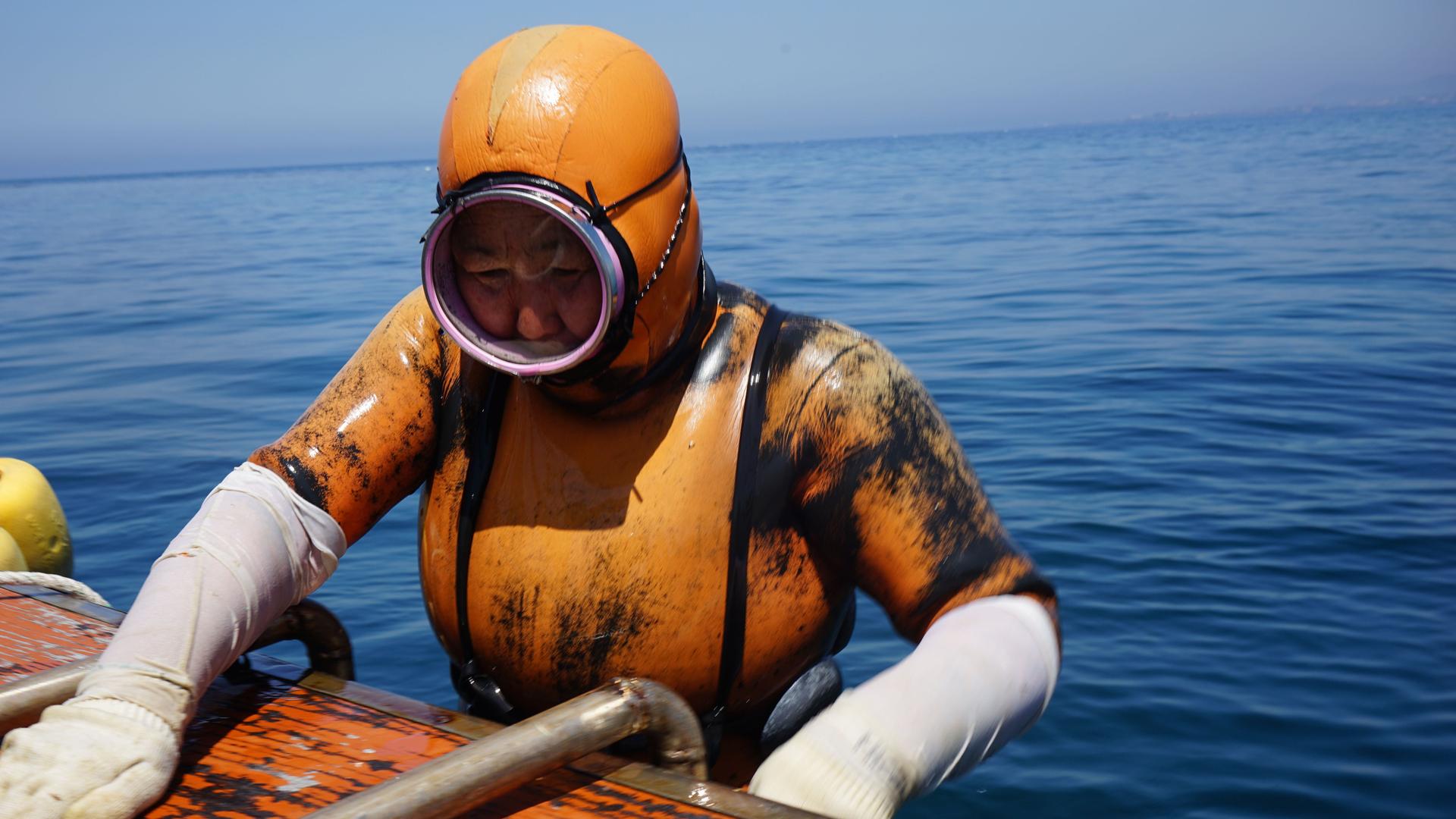 A haeynyo woman climbs aboard the boat. They wear orange diving suits so commercial ships can spot them more easily and steer clear.