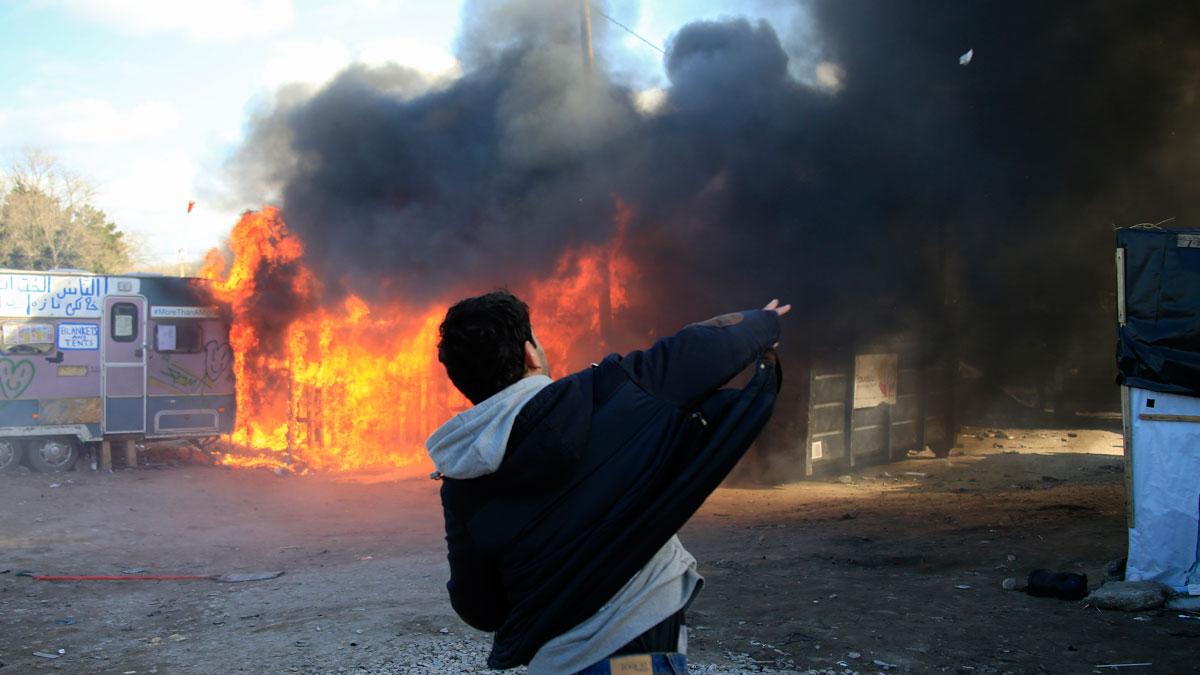 A youth throws a stone as smoke and flames rise from a burning makeshift shelter in protest against the partial dismantlement of the camp for migrants called the "jungle," in Calais.