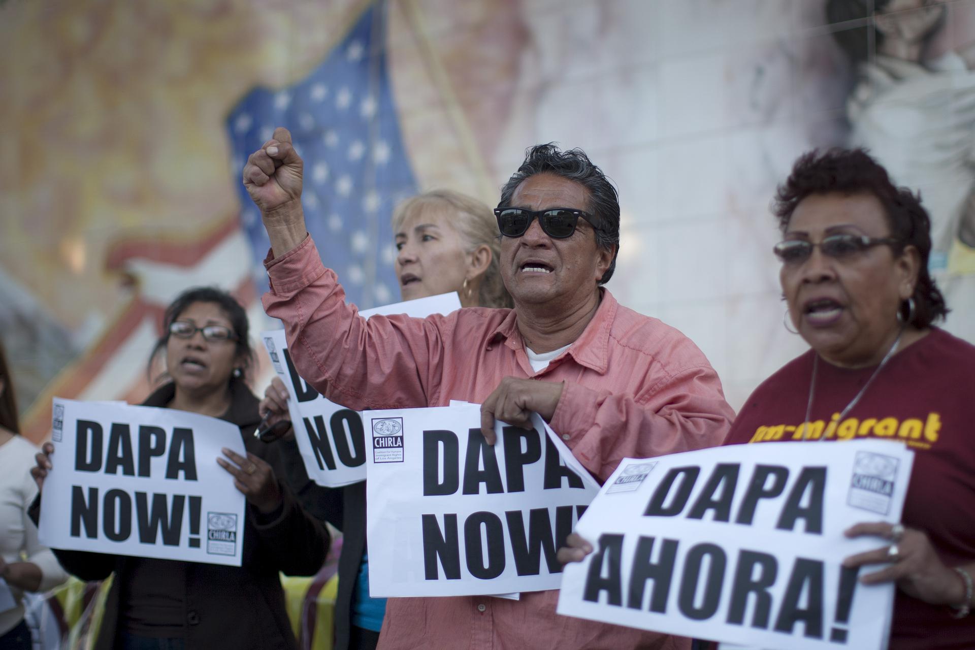 One man and three women hold signs that say "DAPA now!" and "DAPA Ahora!" before a wall covered with a mural, barely visible. 