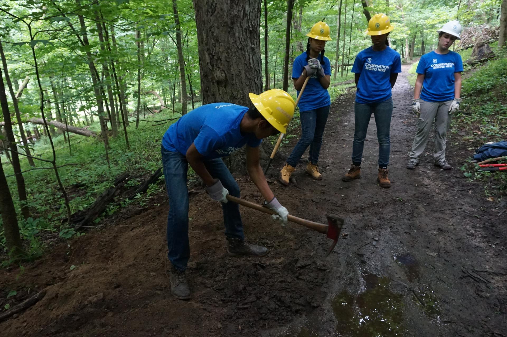 Several young people wearing gloves and helmets work in a park, one digs with a shovel