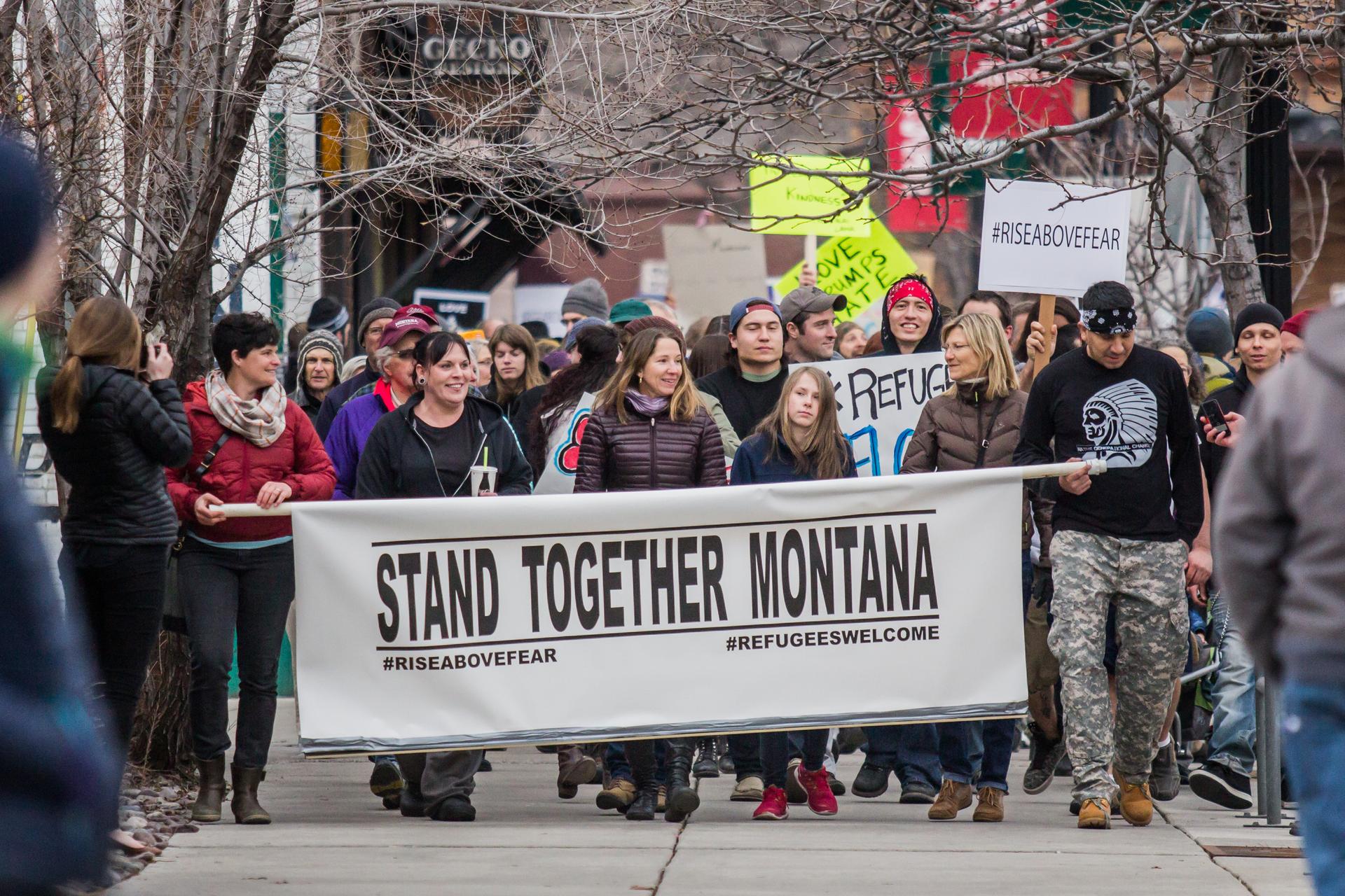 A group of activists march down a street with a large banner that reads, STAND TOGETHER MONTANA.