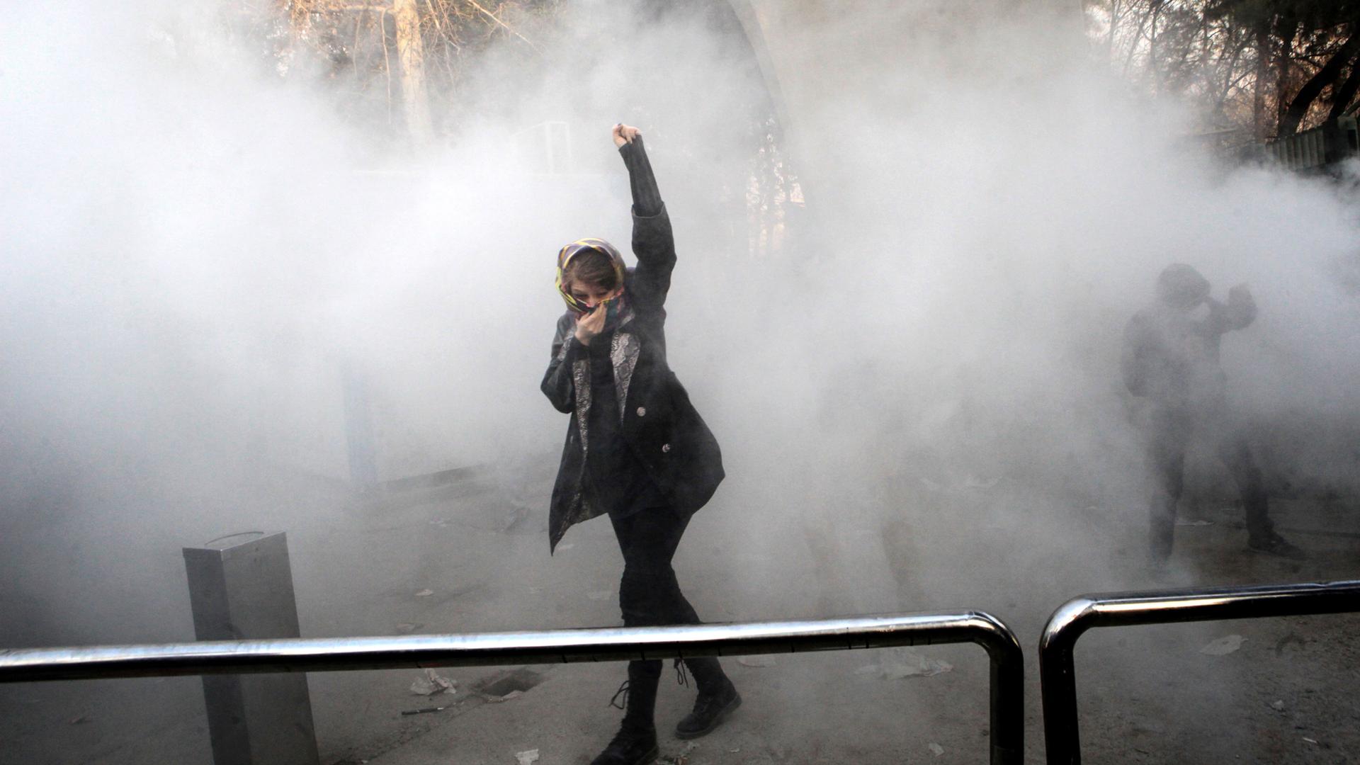An Iranian woman raises her fist, covering her face with a scarf, as smoke surrounds her.