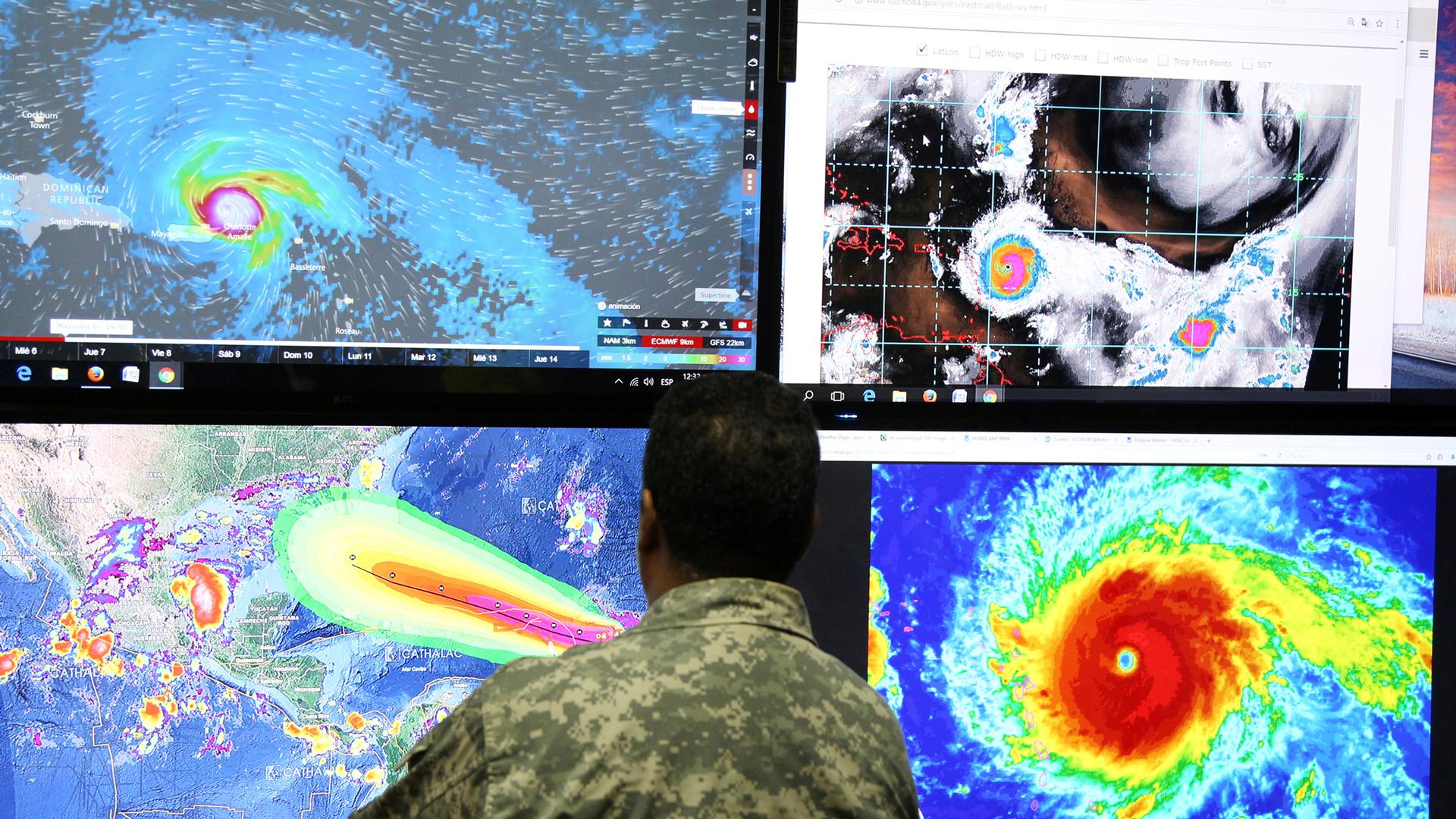 A member of the Emergency Operations Committee standing in front of four TV monitors with images of Hurricane Irma shown.