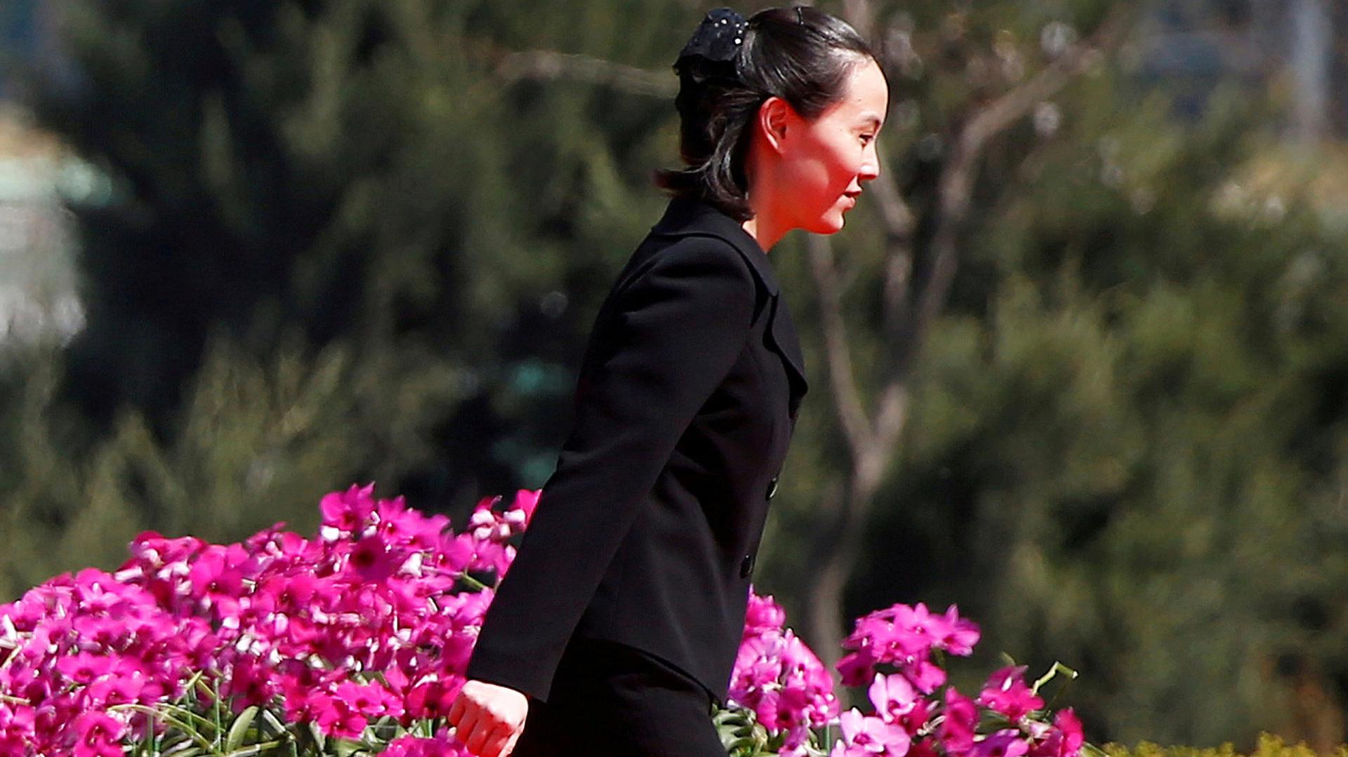 Kim Yo Jong, sister of North Korean leader Kim Jong-un, walks past a flowering bush weaing a black pants suit.