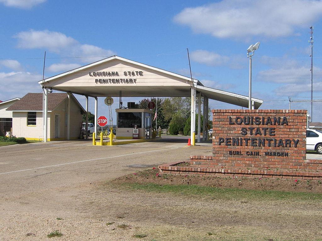 The gate to Louisiana State Penitentiary, pictured here, is a simple two-lane entry and exit way. Before the entrance is a brick wall that says "Louisiana State Penitentiary, Burl Cain, Warden"