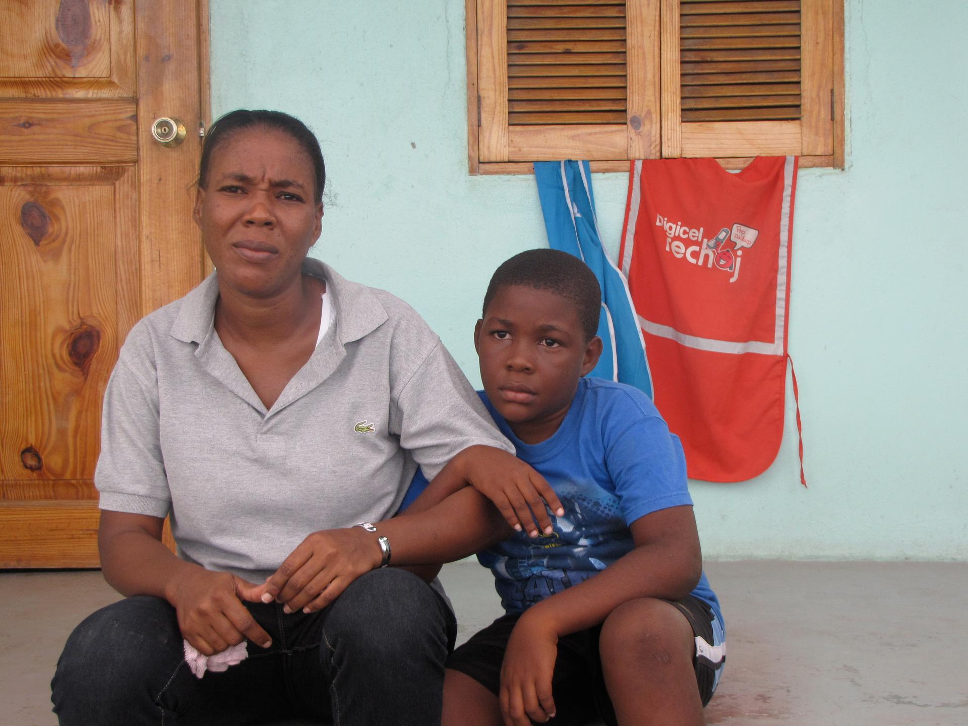 Marie Conce Moreau with her son outside their home in Village la Difference. Moreau took a job at the nearby industrial park when her husband lost his job. They also make ends meet by donning the blue and red aprons to sell phone cards.