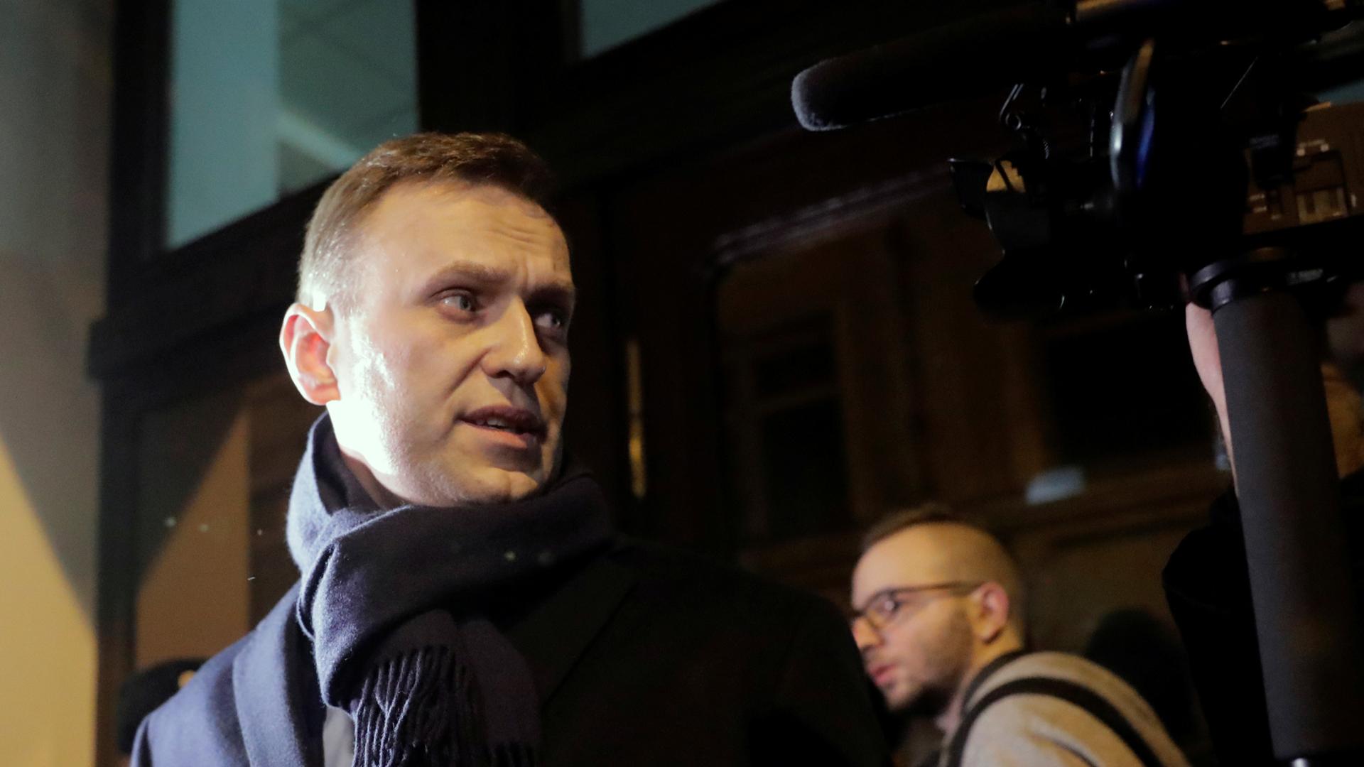 Russian opposition leader Alexei Navalny wearing a dark overcoat and scarf looks left toward a camera.