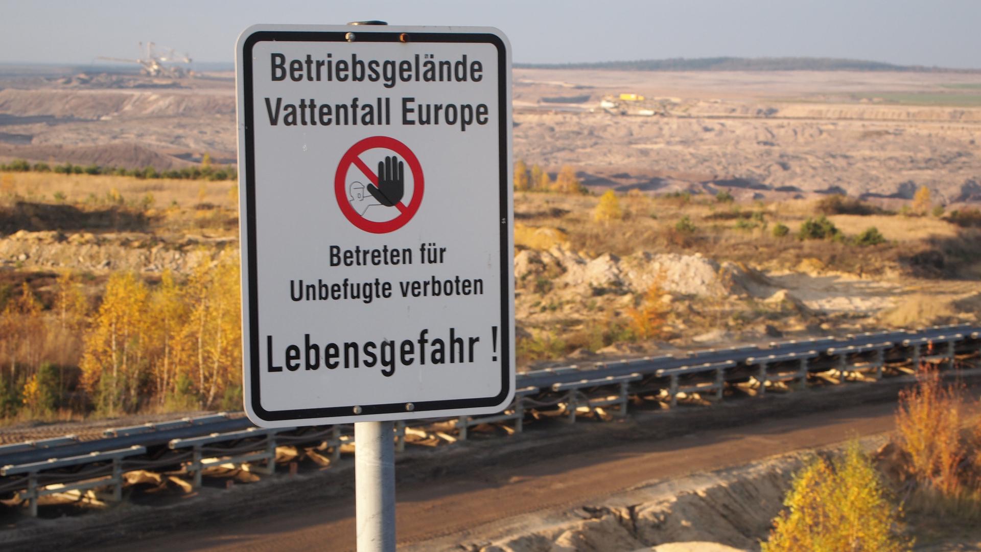 A sign at a German lignite surface mine warns visitors to stay away from the edge of an overlook. A giant earth-moving machine sits on the far horizon.