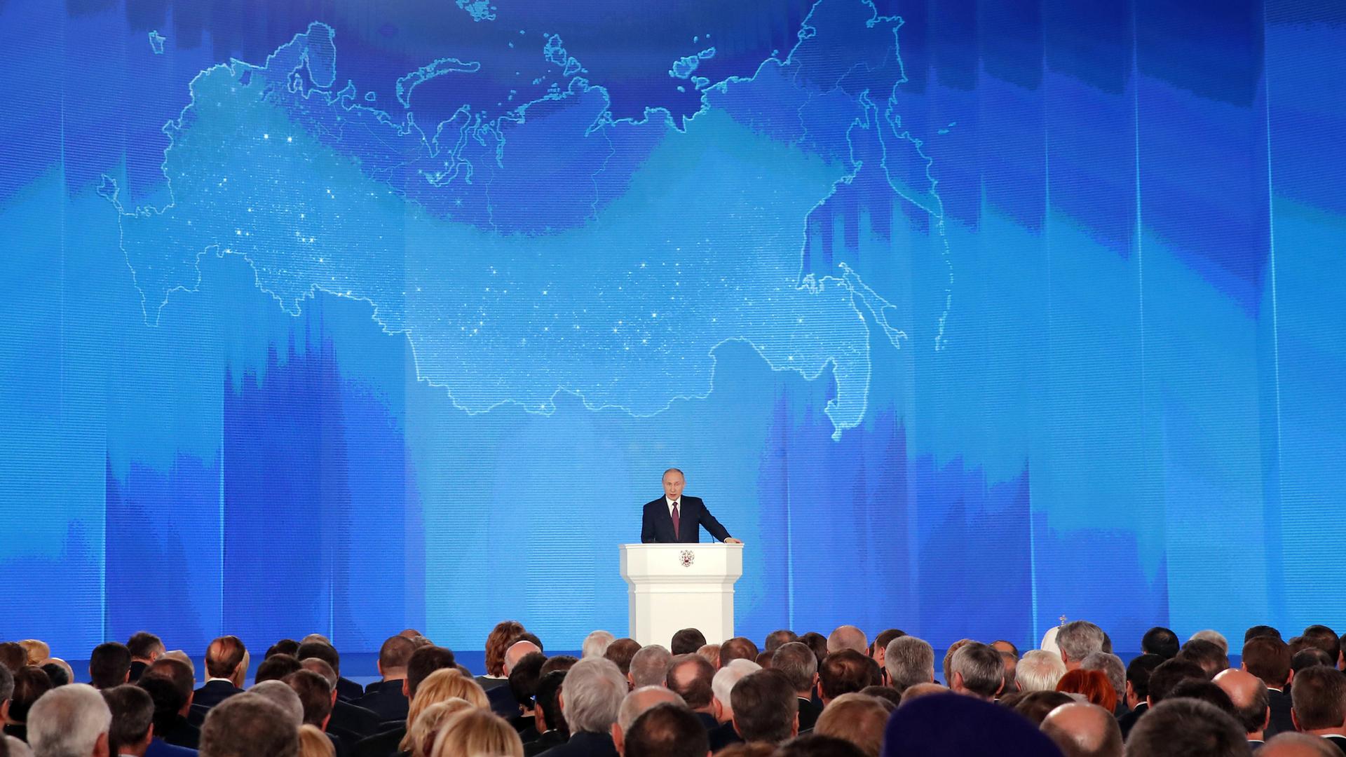 Russian President Vladimir Putin addresses the Federal Assembly in Moscow, standing at a podium in front of a large blue map of Russia.