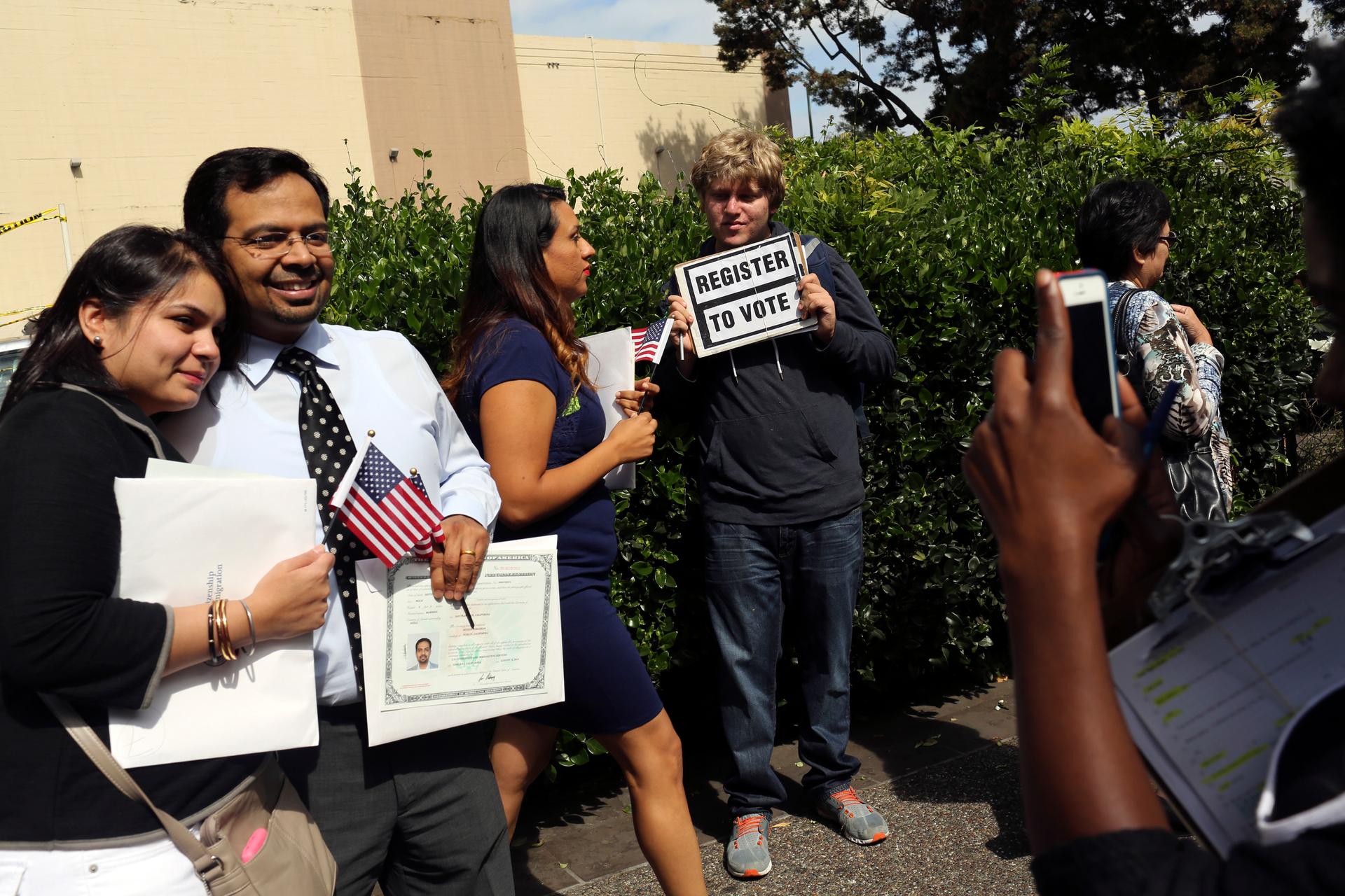 A man holds a voter registration sign while a couple has a souvenir photograph taken following a US Citizenship and Immigration Services ceremony in Oakland, Californiam on August 13, 2014. 