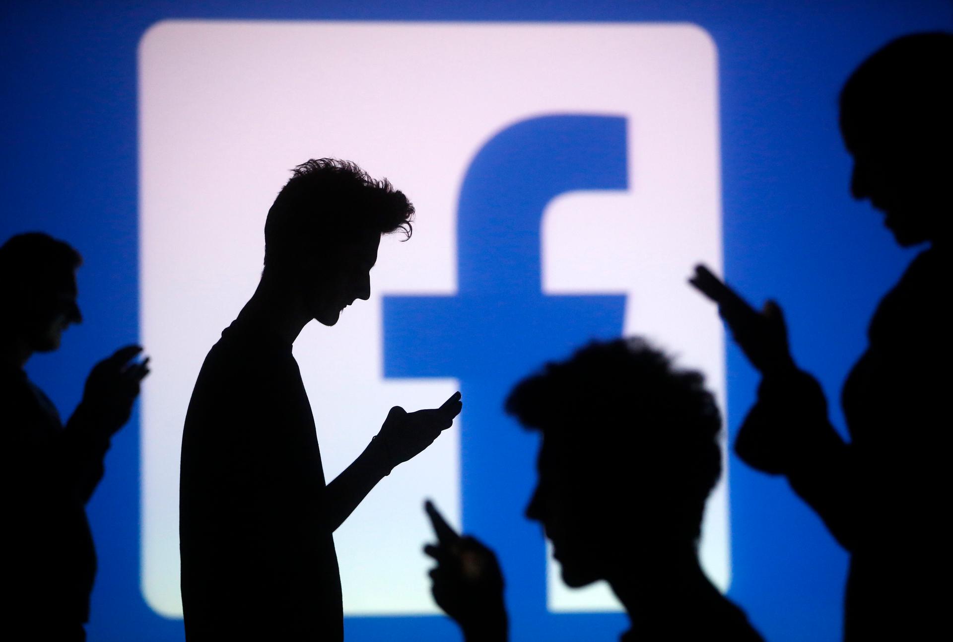 People are silhouetted as they pose with mobile devices in front of a screen projected with a Facebook logo