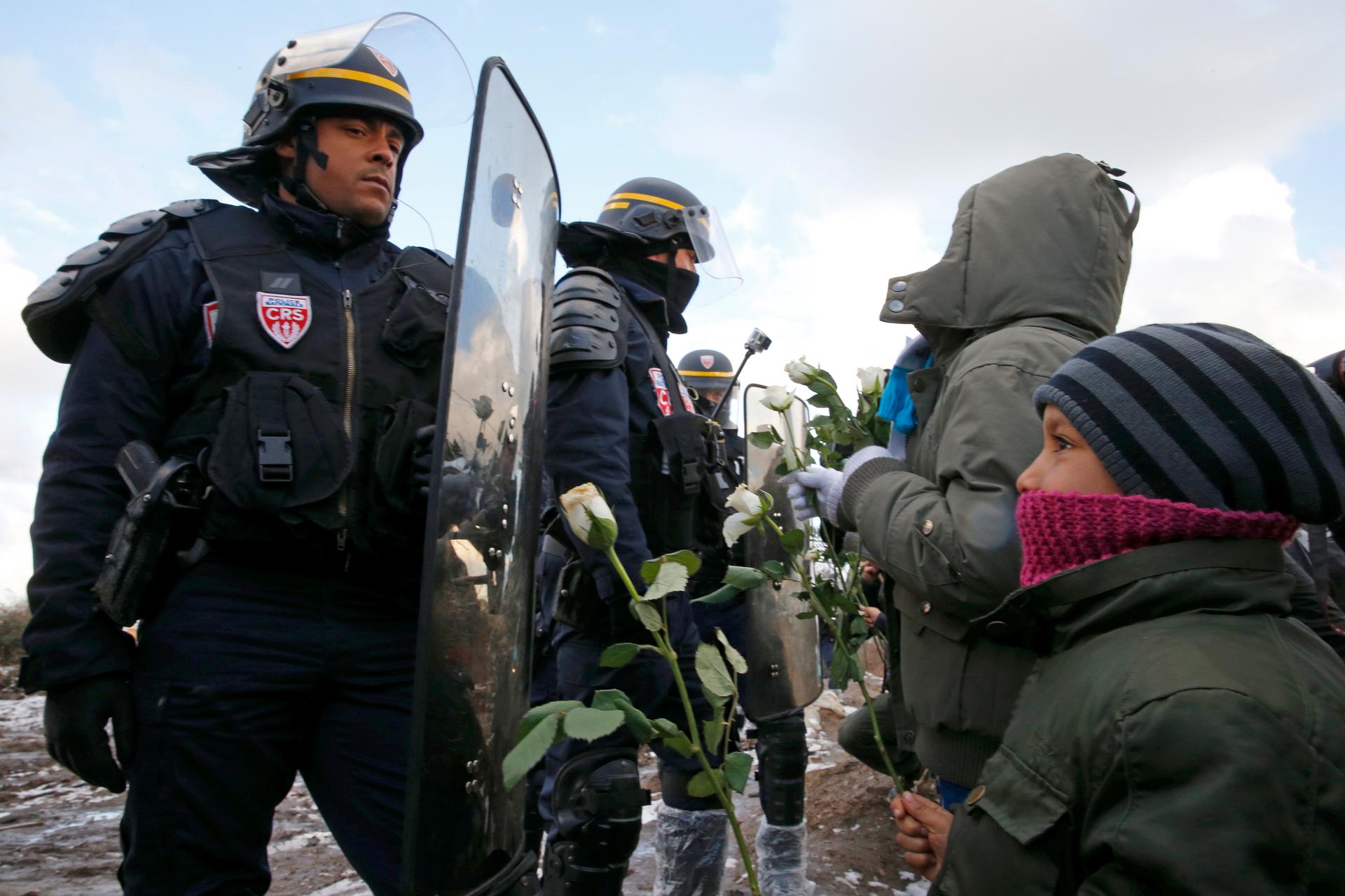 Youngs migrants holding white flowers face French riot police officers who secure the area near makeshift shelters during the partial dismantlement of the camp for migrants called the "Jungle" in Calais, France, March 7, 2016.