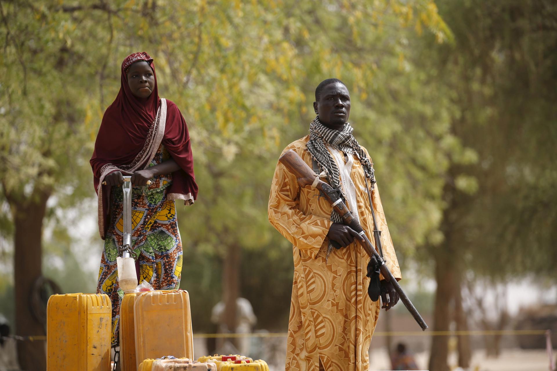 A member of a civilian vigilante group holds a hunting rifle while a woman pumps water into jerrycans in Kerawa, Cameroon, March 16, 2016.