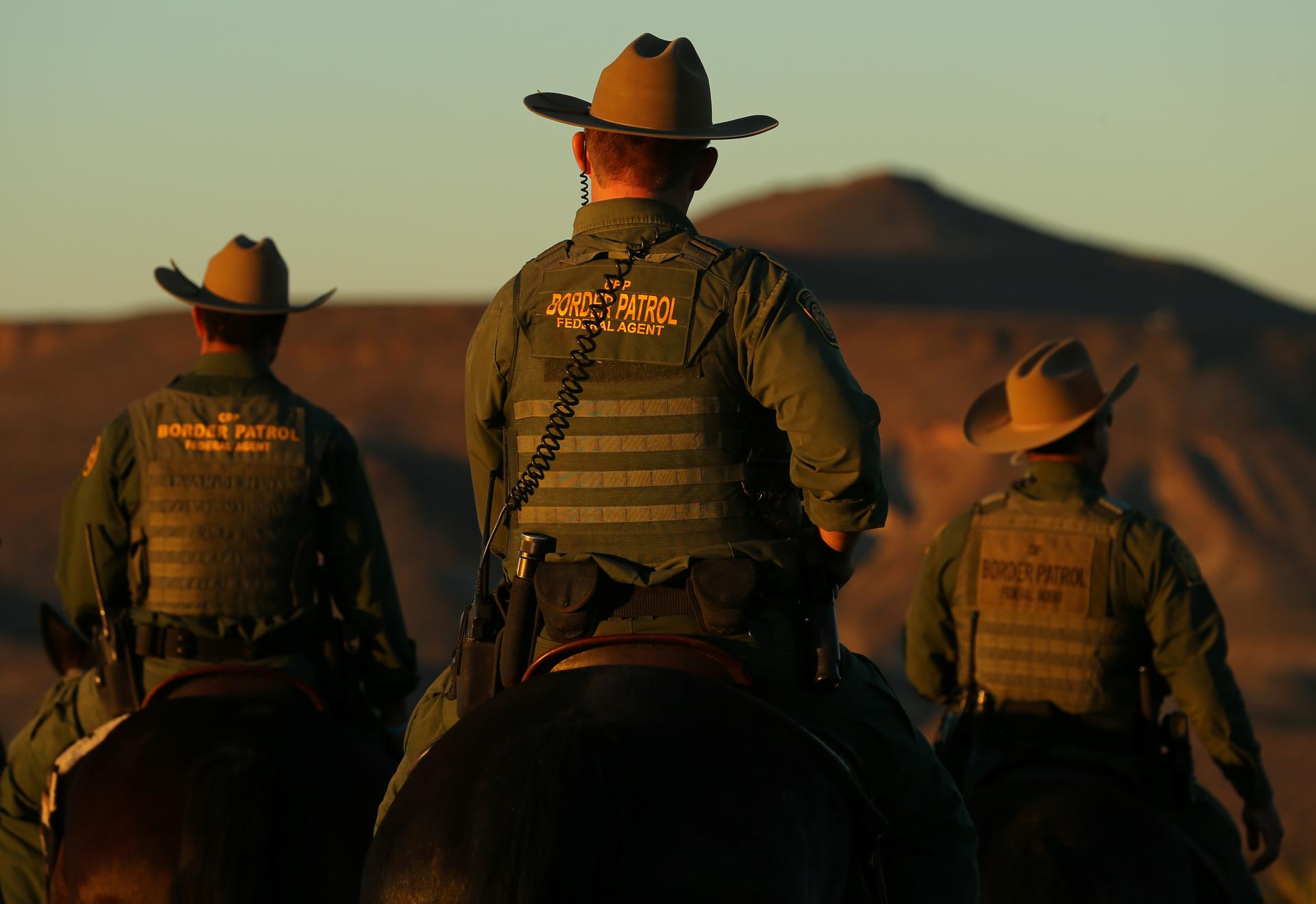 Men on horses in Border Patrol uniforms, seen from back at dusk