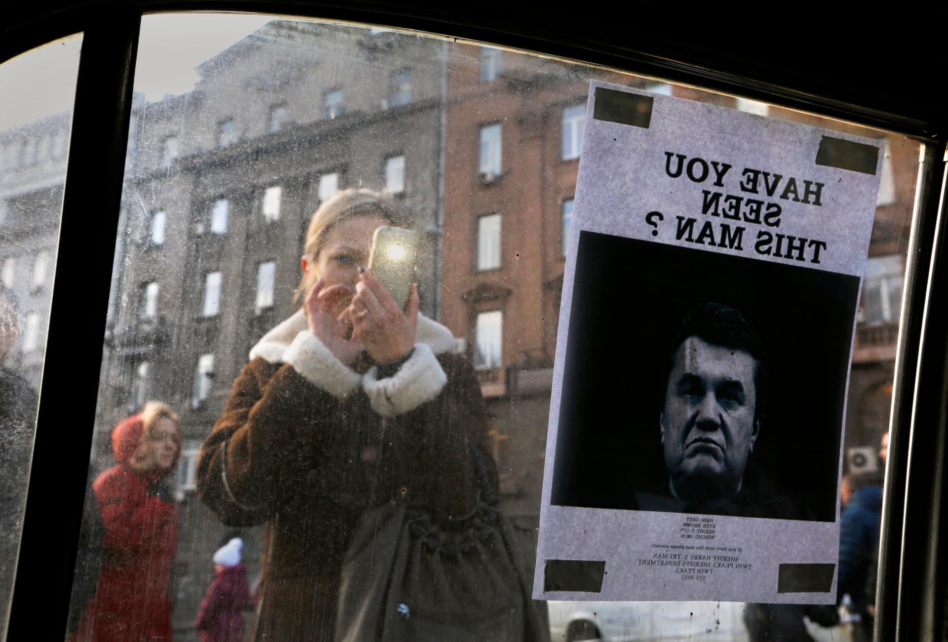 A woman takes photos of a "Wanted" notice for fugitive Ukrainian President Victor Yanukovich, plastered on the window of a car used as a barricade, near Kiev's Independent Square February 24, 2014. 
