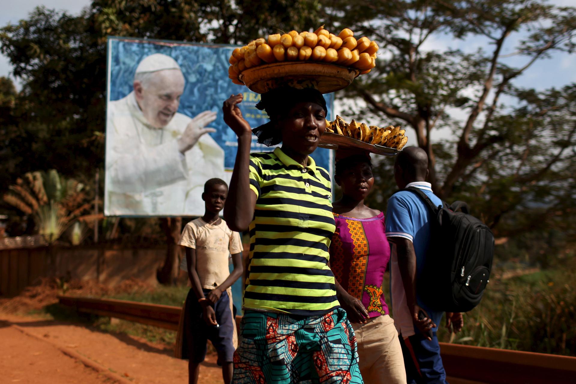Women carrying fruits and vegetables on their heads walk past a billboard with a photograph of Pope Francis, in Bangui, Central African Republic; November 26, 2015.