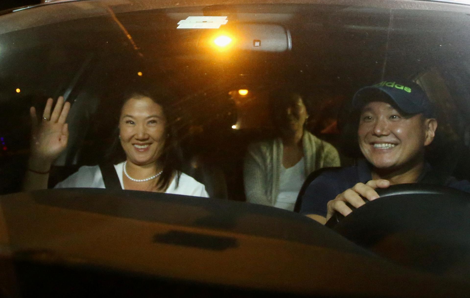 three people inside car, man driving, woman in passenger seat smiling and waving, one more person in back