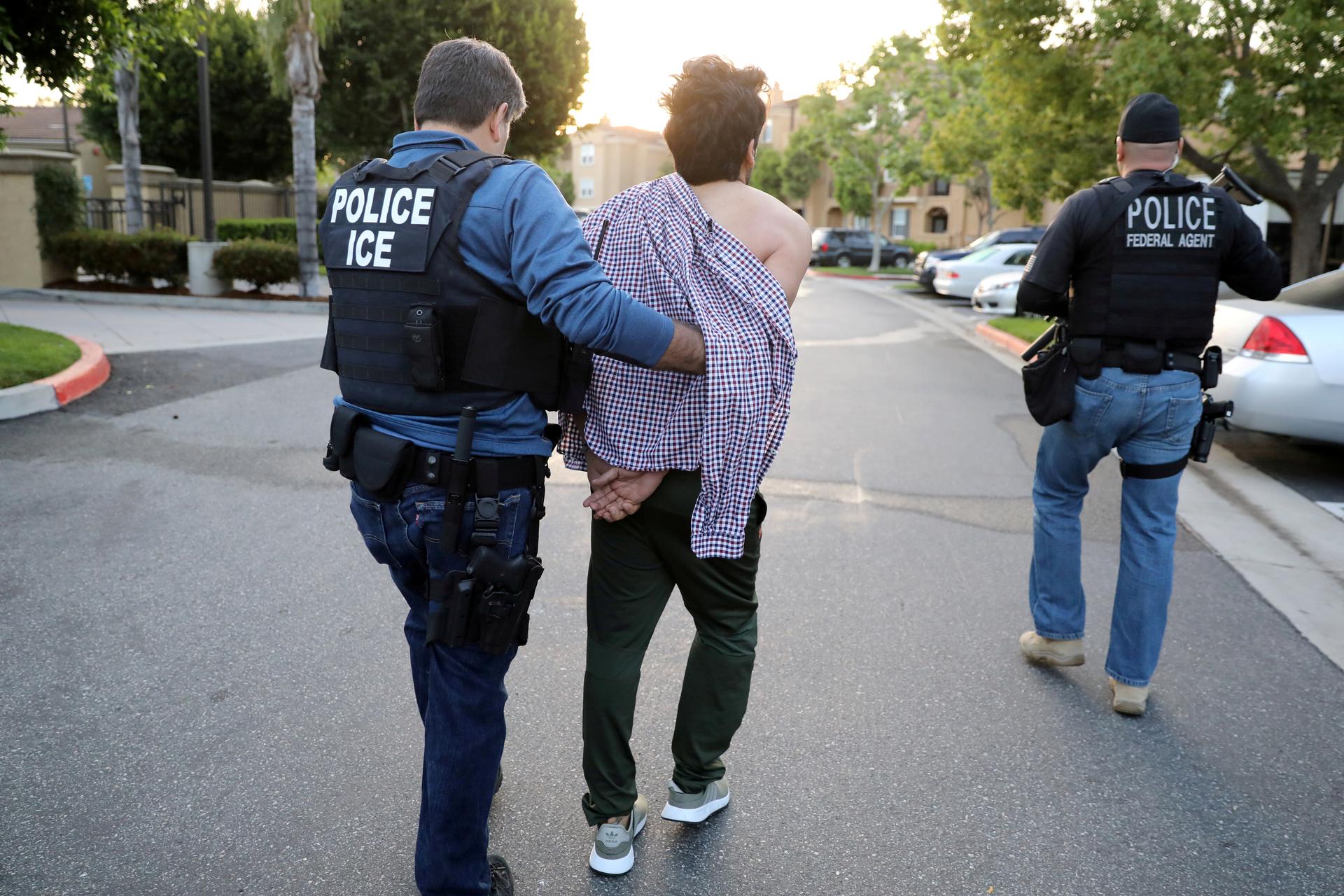 A man with "POLICE ICE" protective vest leads a man in handcuffs down street, with another officer with "POLICE FEDERAL AGENT" on vest on the right