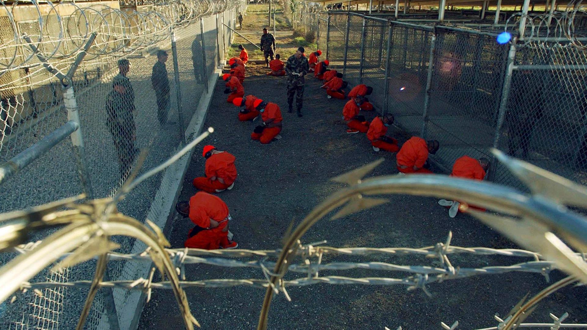 Detainees in orange jumpsuits sit in a holding area under the watchful eyes of military police during in-processing to the temporary detention facility at Naval Base Guantanamo Bay in January 2002.