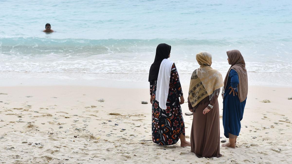 Three women in hijab wait as a boy swims in the ocean in front of the Casa Nemo Beach Resort and Spa of Pulau Weh island, Indonesia.