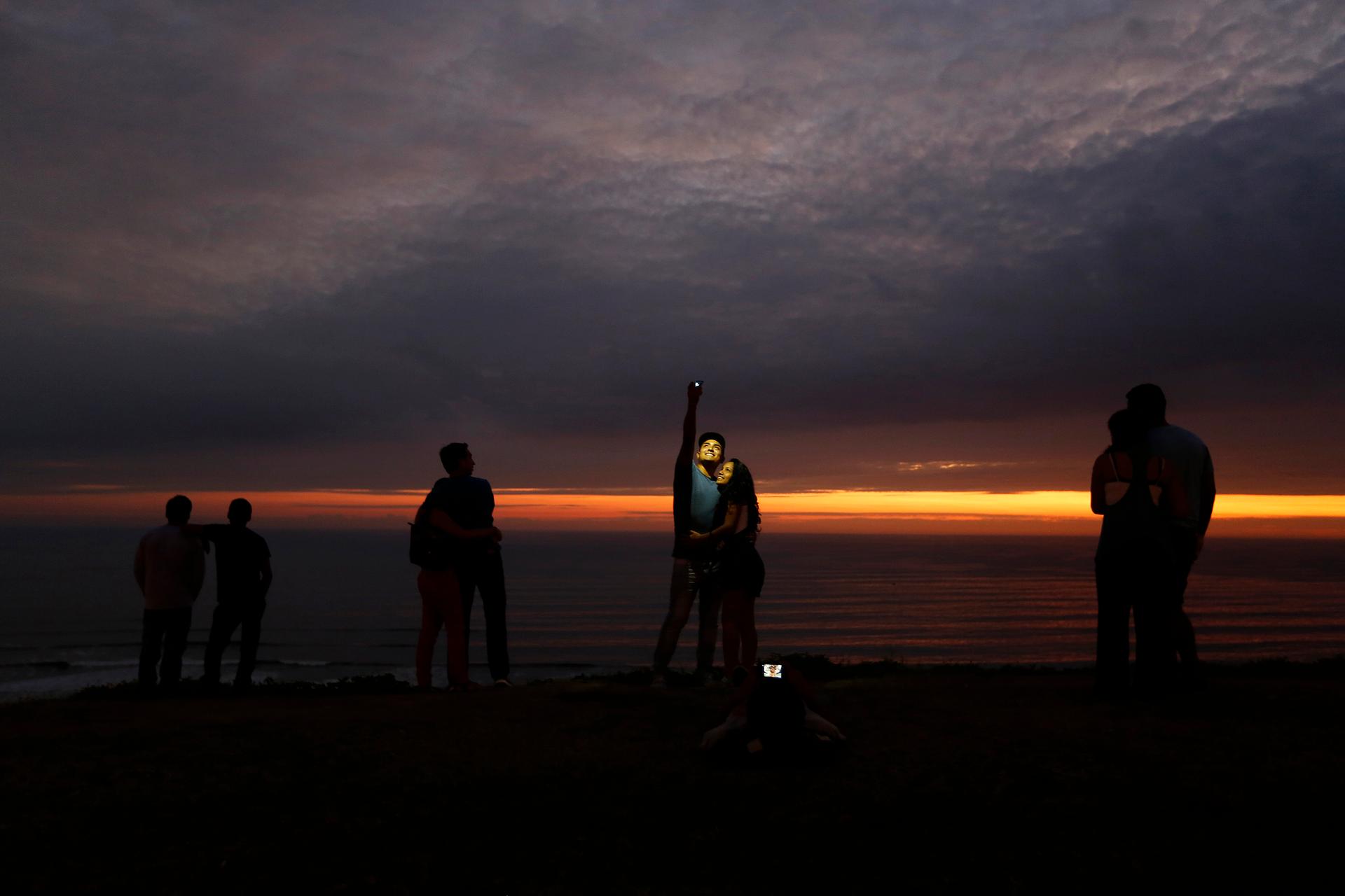 A couple takes a selfie as others observe the sunset at an oceanfront in the neighborhood of Miraflores in Lima.