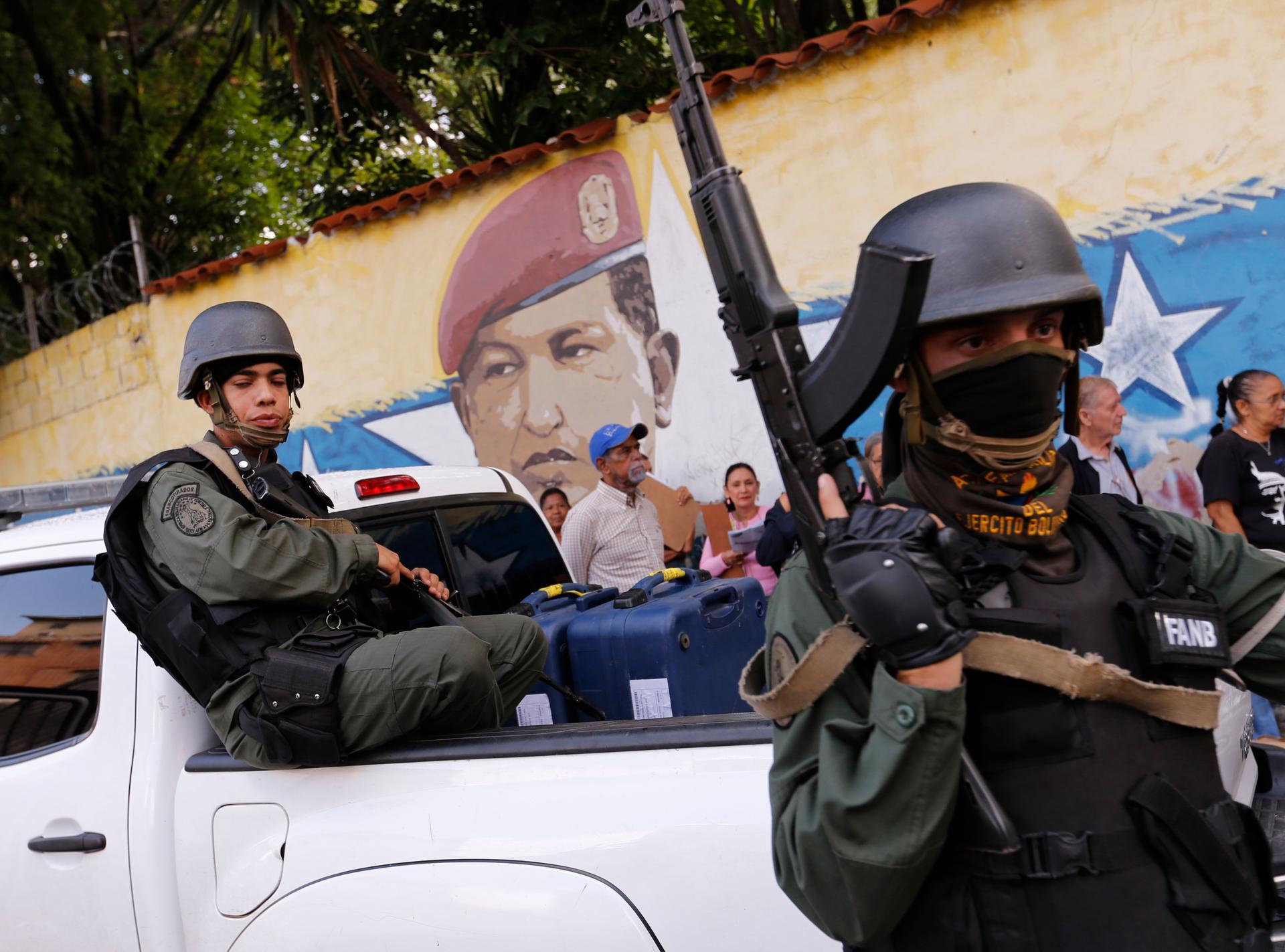 One soldier sits in a truck and another stands with riffle in hand in front of a mural of the late Venezuelan President Hugo Chavez.