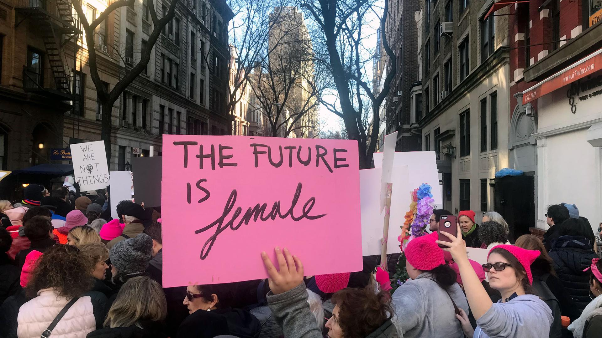 A pink sign reading, "The future is female" is hoisted above the crowds of marchers, some of whom are wearing the signature pink pussy hats.