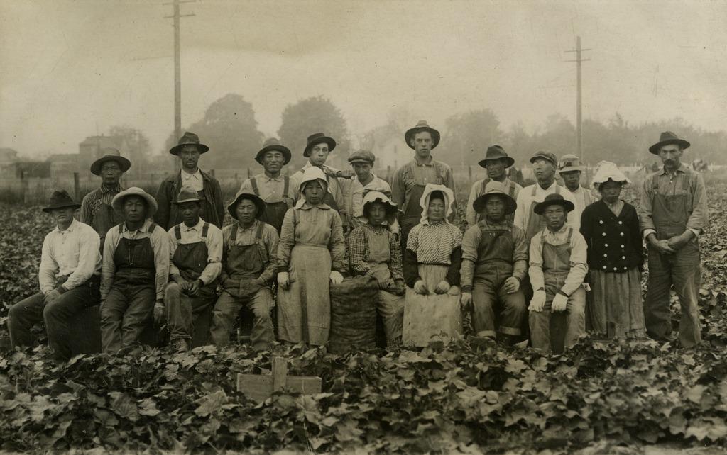 A group of workers pose in a farm, wearing their work clothes. Top row standing, bottom row sitting. Black and white photo.