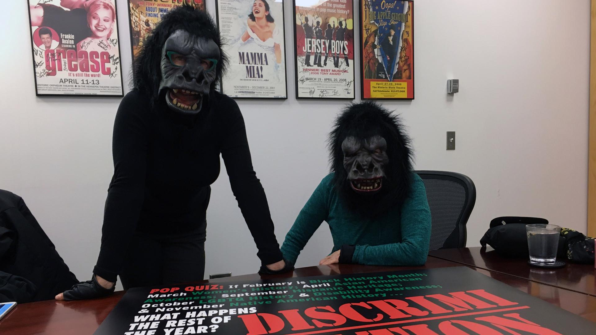 Two women in gorilla masks pose in front of their poster, which critiques the special months devoted to the history of marginalized groups.