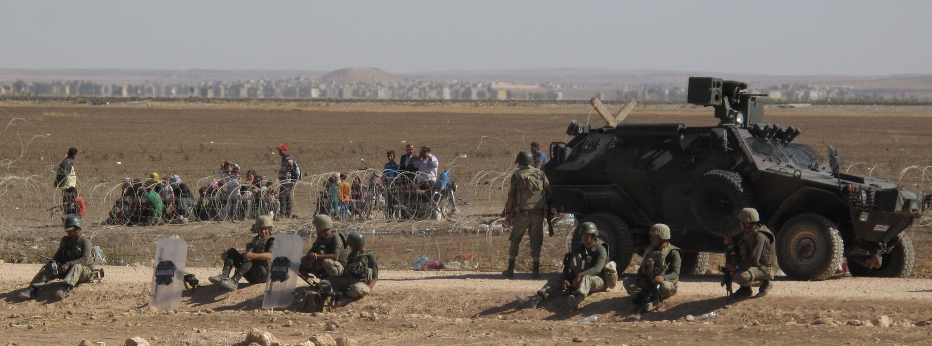 Scores of Syrian refugees from Kobane - under threat of capture by Islamic State - sit in the sun waiting to be allowed to cross into Turkey. Sept. 26, 2014. The outskirts of the village are visible in the background.