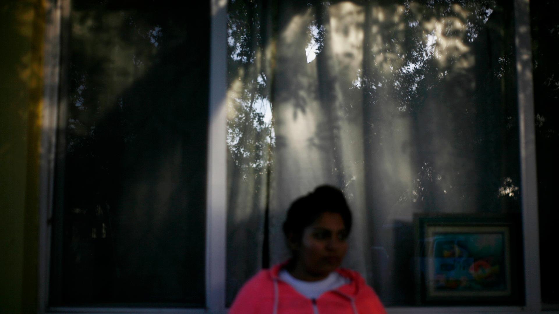 Blurry image of woman in pink hodded sweatshirt standing in front of window, with reflection of trees