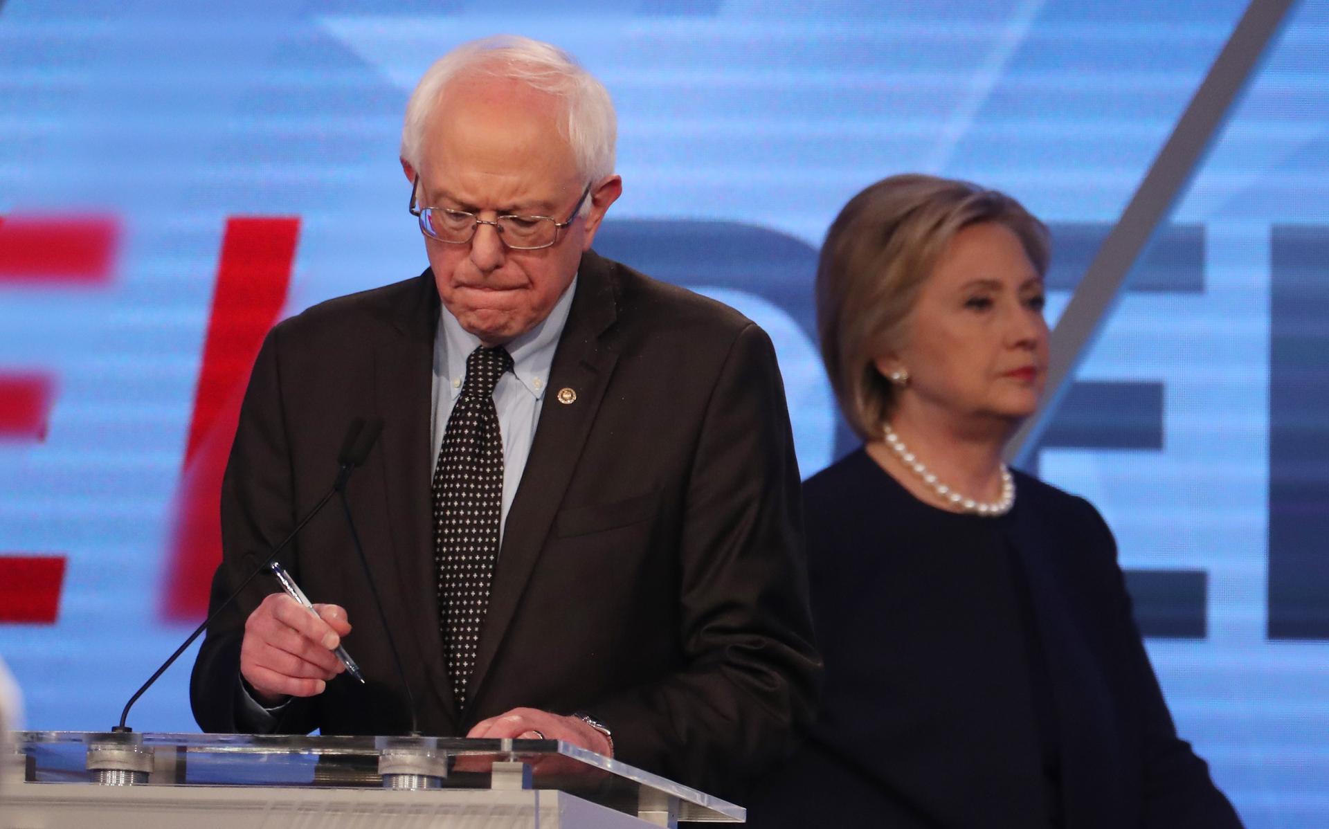 Democratic US presidential candidate Senator Bernie Sanders writes on his notes as his rival Hillary Clinton walks behind him