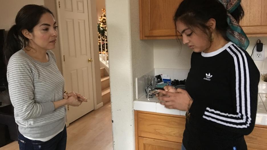 Two woman stand in kitchen, one older and one younger looking at her phone