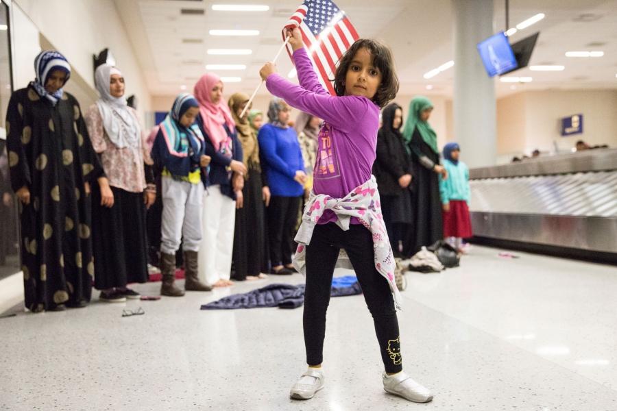 A young girl dances with an American flag in baggage claim while women pray behind her during a protest at the Dallas/Fort Worth International Airport.