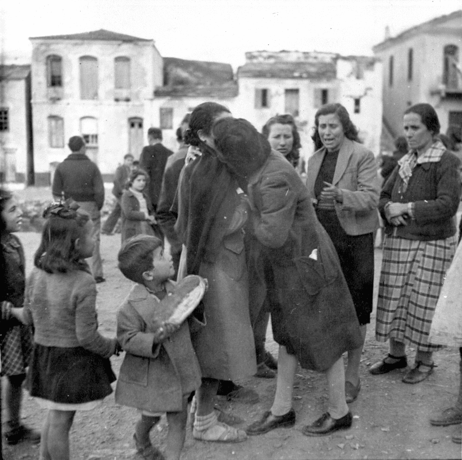 Two women hug while children look on, and other women look on crying. In black and white