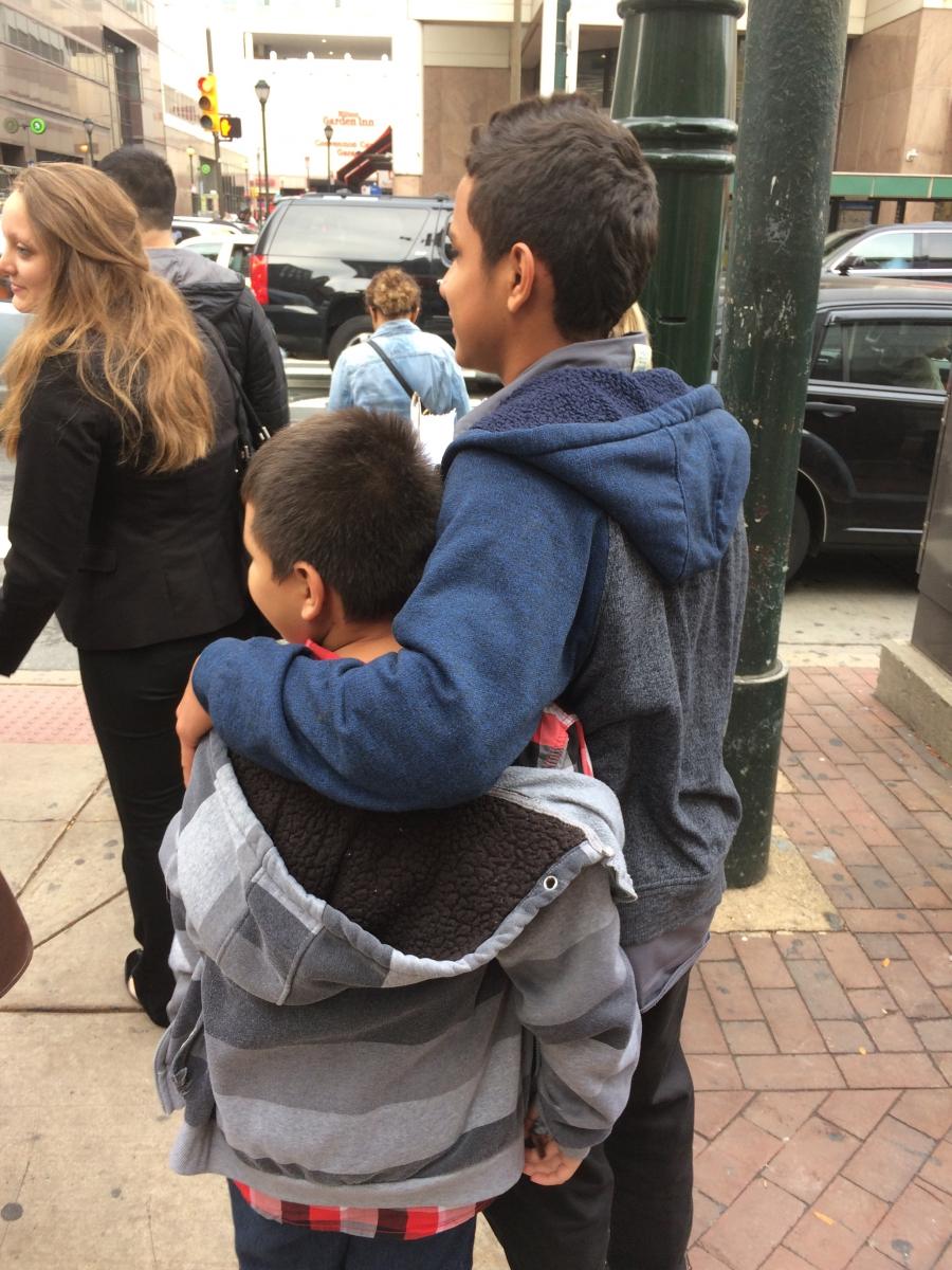 Two young boys face away from camera, on sidewalk