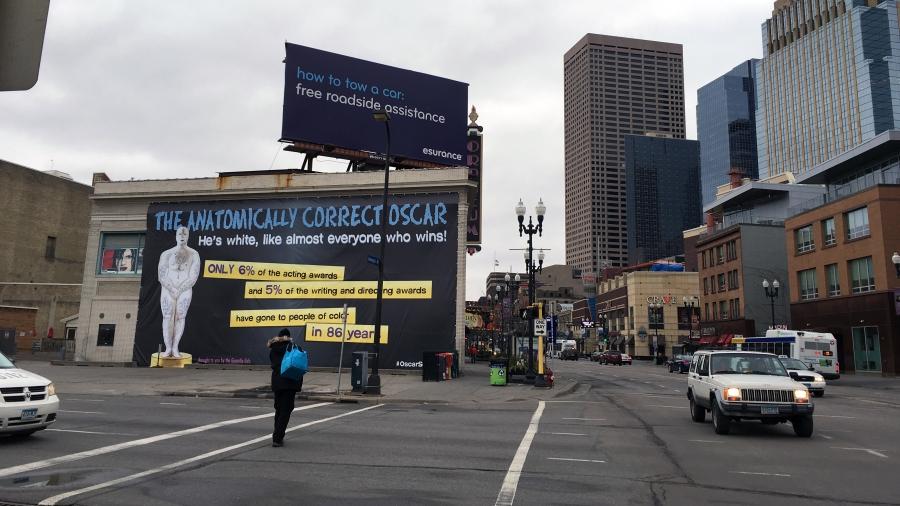 A view of a Guerrilla Girls poster in Minneapolis from across the street. The poster shows a cartoon version of the Oscar as a white man and covers one side of a midrise building downtown.